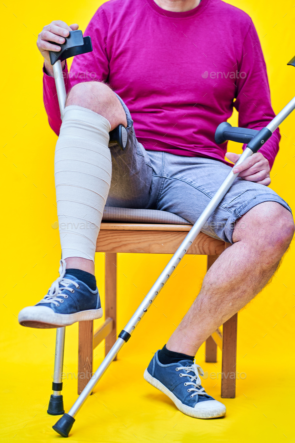 Man with crutches sitting on a chair, supporting a bandaged leg on the
